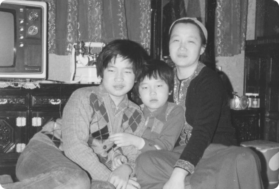 A family of three, two older siblings and a young child, pose together in a home living room with a vintage television set in a black-and-white photograph from the 1970s or 1980s.