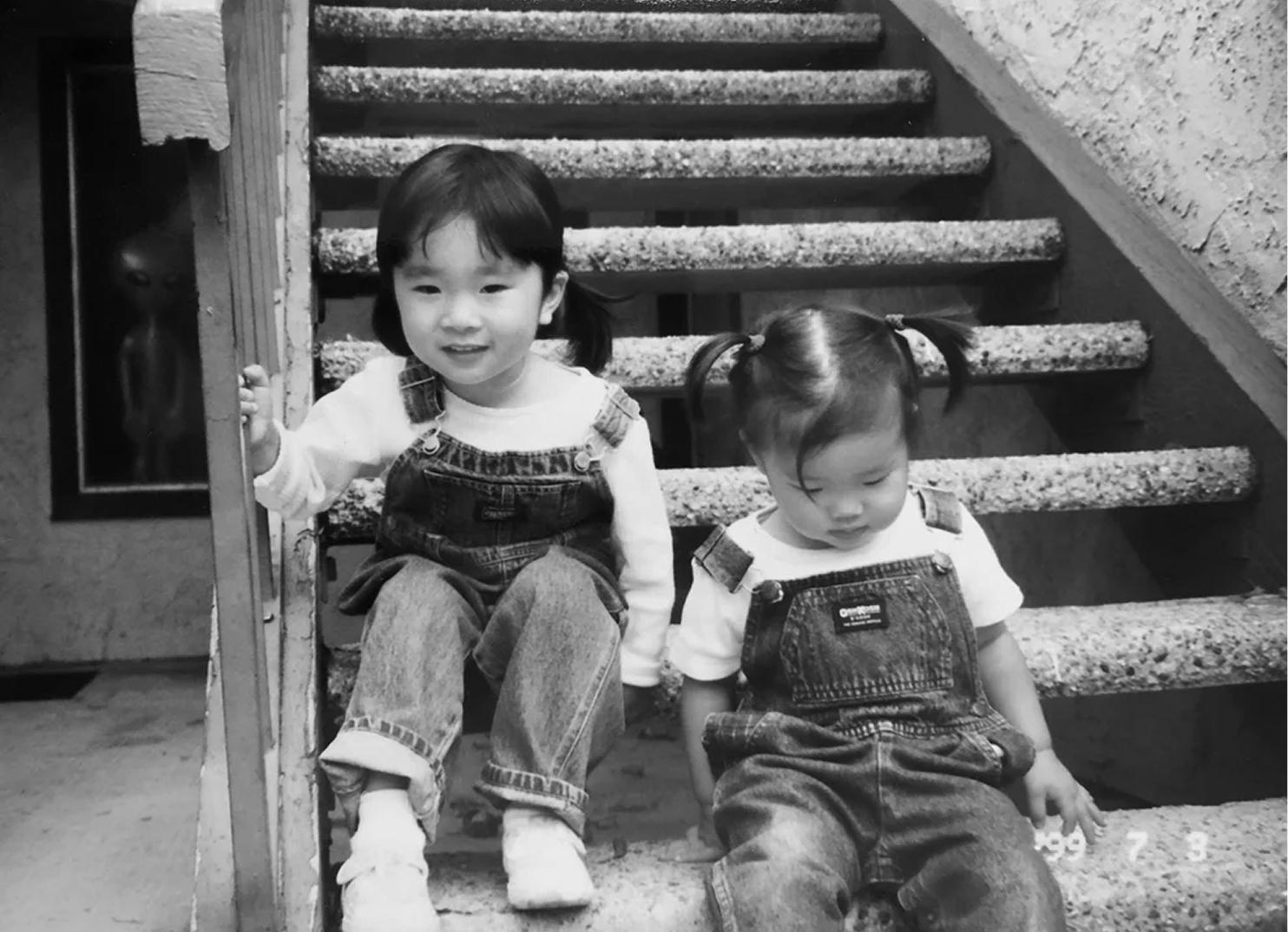 Two young girls in matching denim overalls sit together on outdoor concrete steps in a black-and-white photograph.