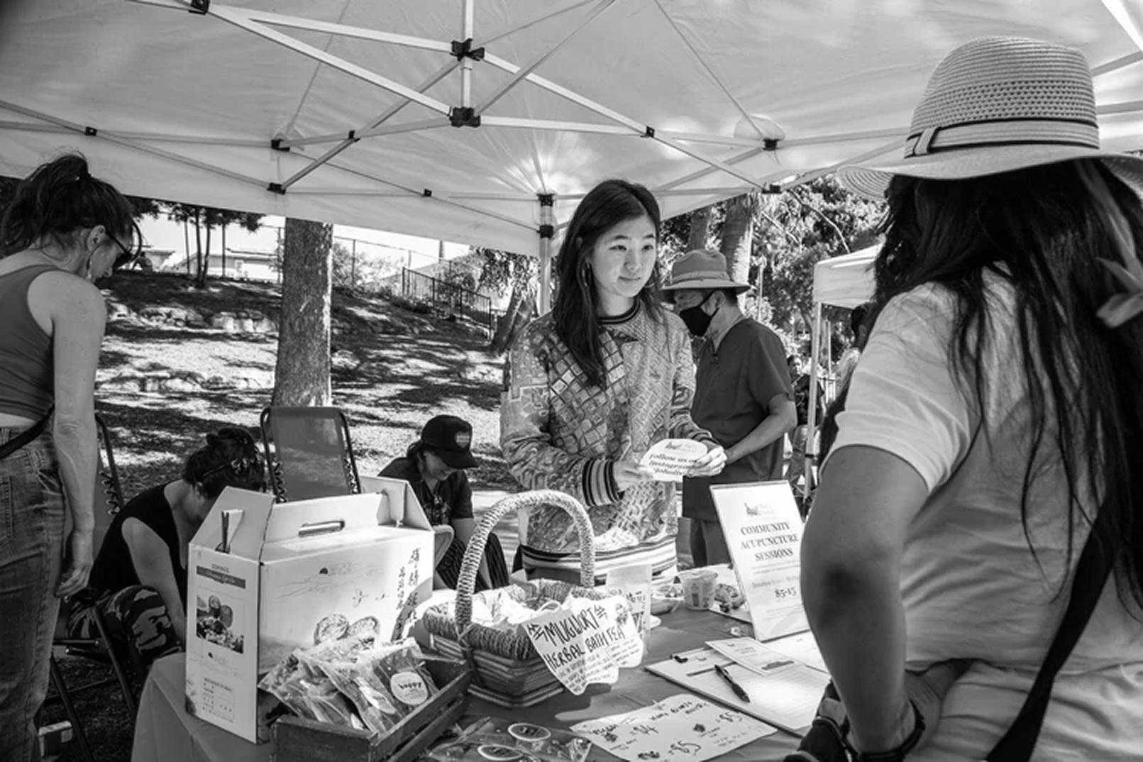A woman staffs an outdoor community health booth under a white canopy tent offering herbal bath tea and community acupuncture sessions while speaking with an attendee at an outdoor event.