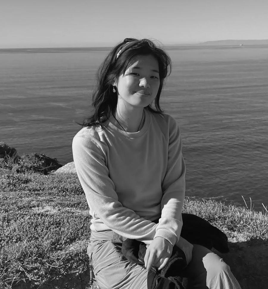A young woman sits on a grassy coastal bluff with the ocean and horizon visible behind her.