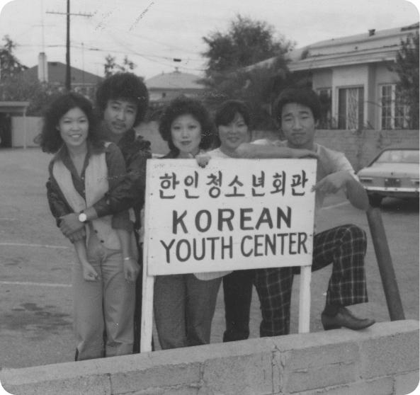 Pedestrians crossing a Koreatown street past parked cars and Korean-language shop signs, photographed in black and white.