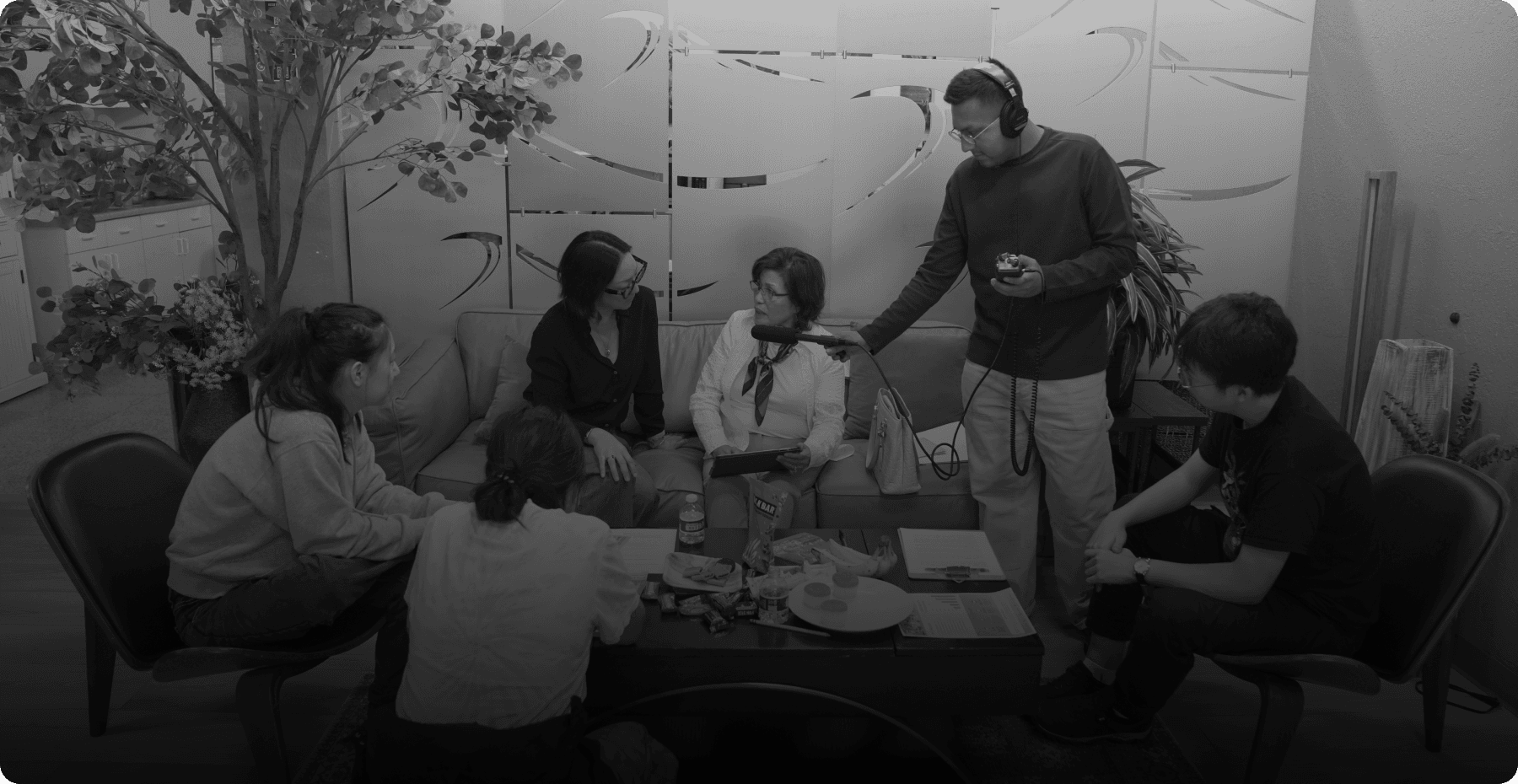 A journalist wearing headphones holds a microphone toward a woman seated on a couch during an interview while several other people look on in an indoor setting with decorative frosted glass panels.