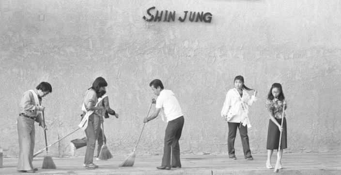 A line of people in white shirts work together clearing a paved lot in front of a wall painted with large lettering, in a black-and-white photograph.