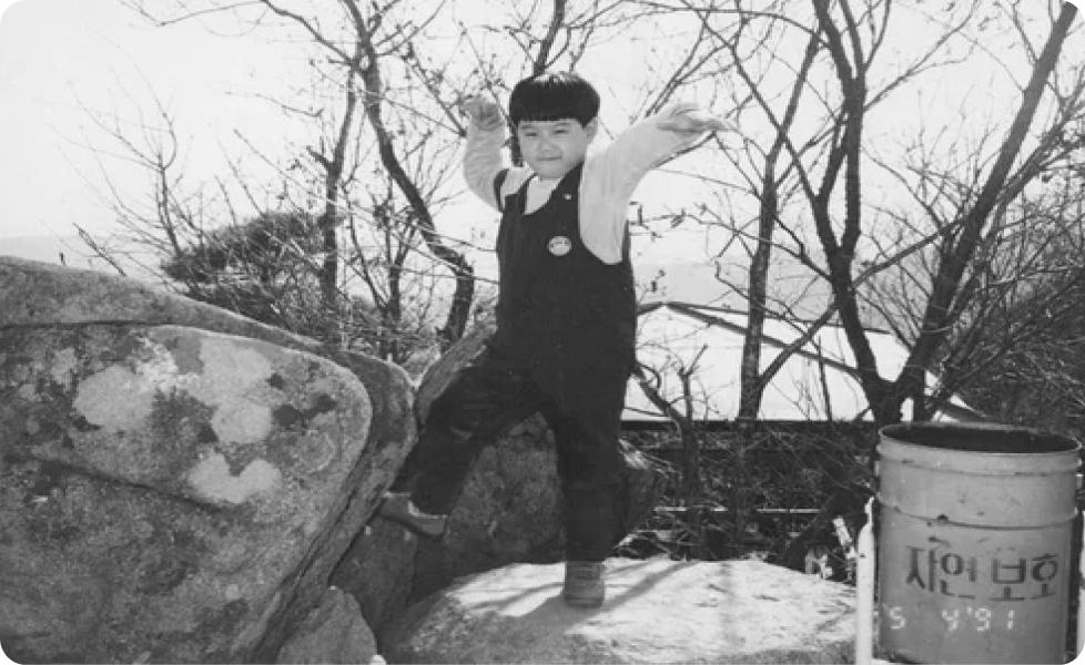 A young boy strikes a playful martial arts pose on a large rock outdoors in a black-and-white photograph with bare winter trees and a Korean-language trash can visible in the background.