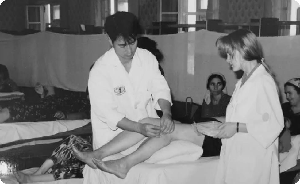 A practitioner in a white coat performs acupuncture on a patient's leg while a colleague assists in a clinical setting with other patients visible in the background.