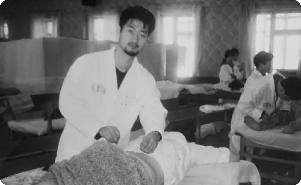 A practitioner in a white lab coat treats a patient lying on a table in a busy clinic ward with other staff and patients visible in the background.