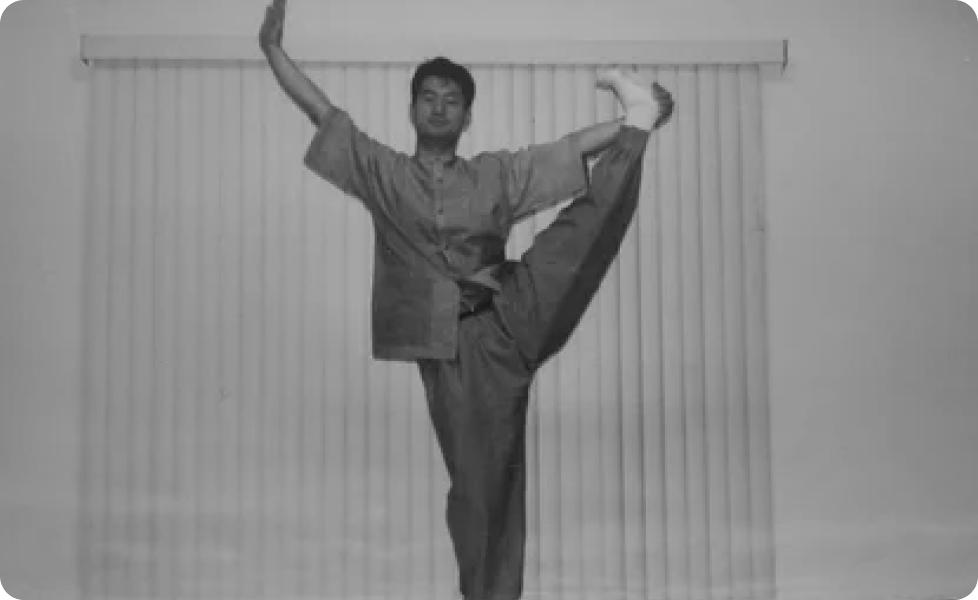 A martial artist in traditional uniform performs a high kick with one arm raised in a balanced stance in front of vertical blinds.