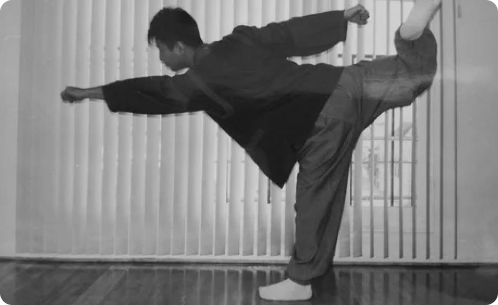 A martial artist in a dark uniform executes a powerful side kick leaning forward with one arm extended in front of vertical blinds on a hardwood floor.