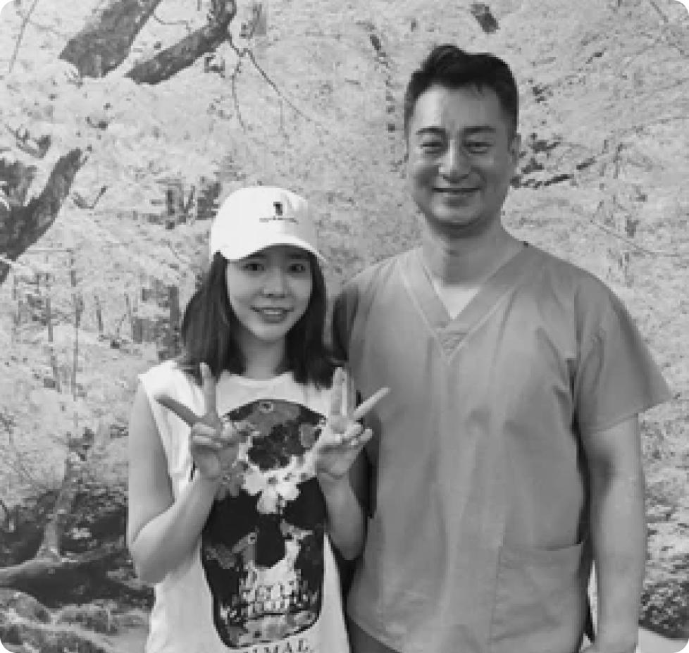 Two people pose together making peace signs in front of a blossoming tree backdrop; one wears a baseball cap and graphic tee and the other wears medical scrubs.