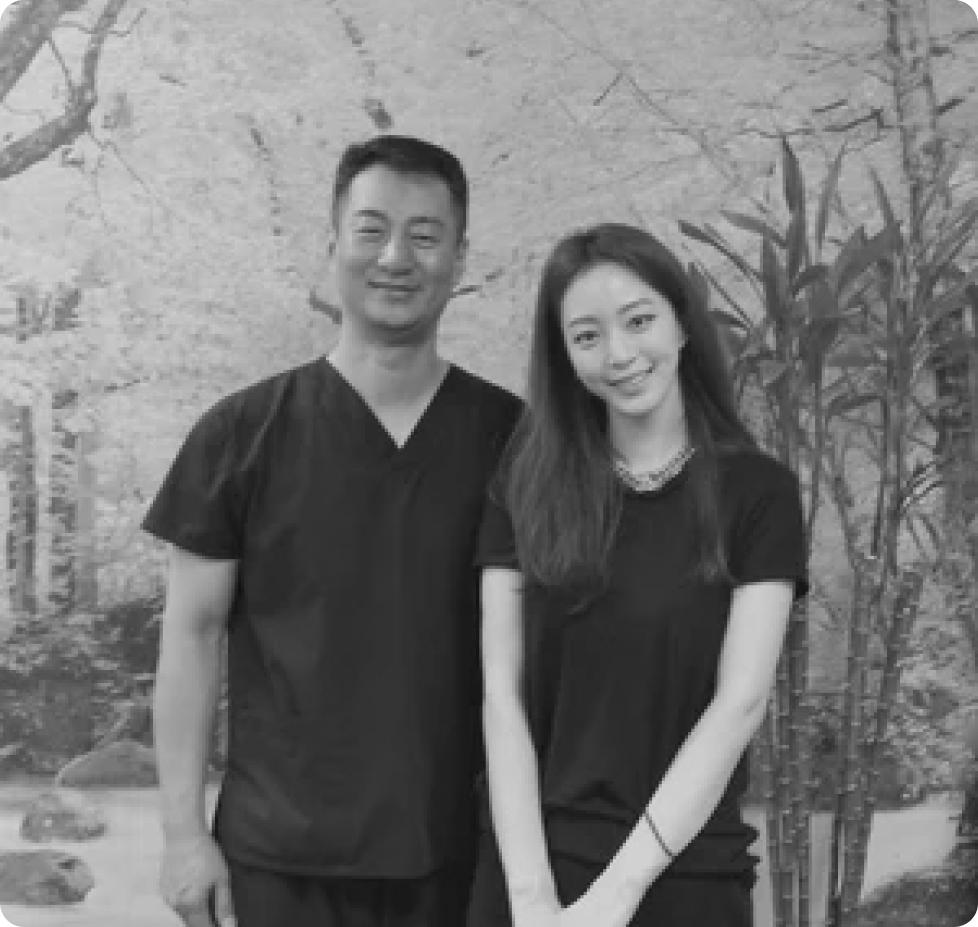Two people in matching dark scrubs smile together in front of a serene garden backdrop with bamboo and flowering trees.