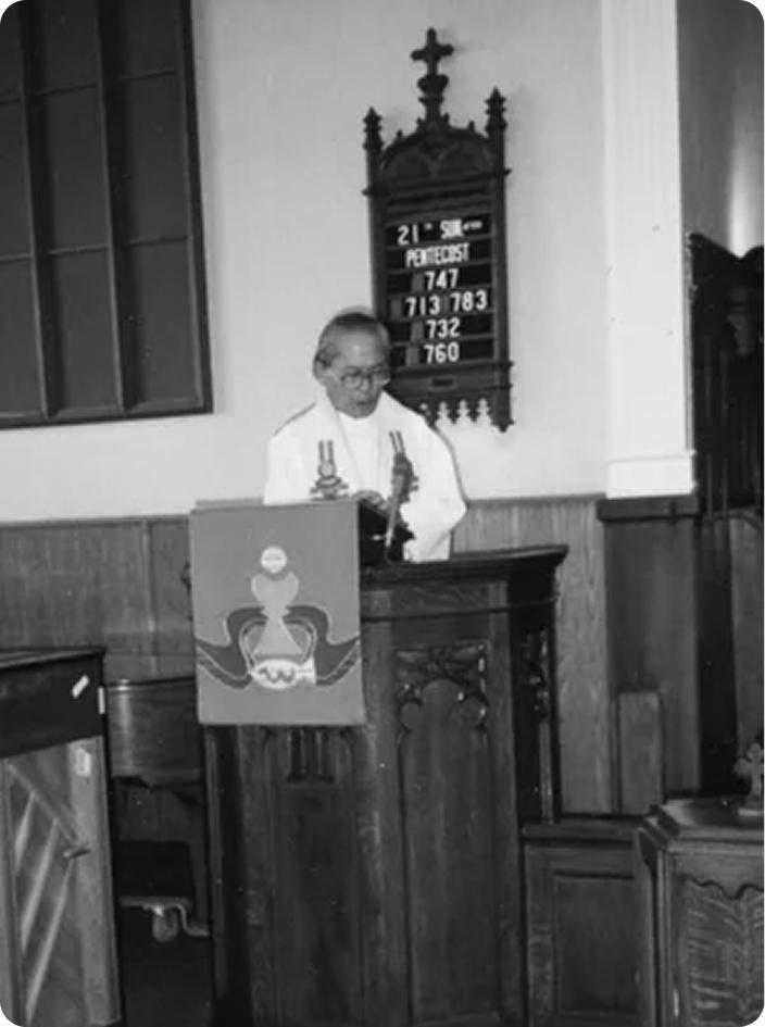 A minister in clerical robes delivers a sermon at a wooden pulpit inside a church with a hymn board showing the 21st Sunday of Pentecost visible on the wall behind him.