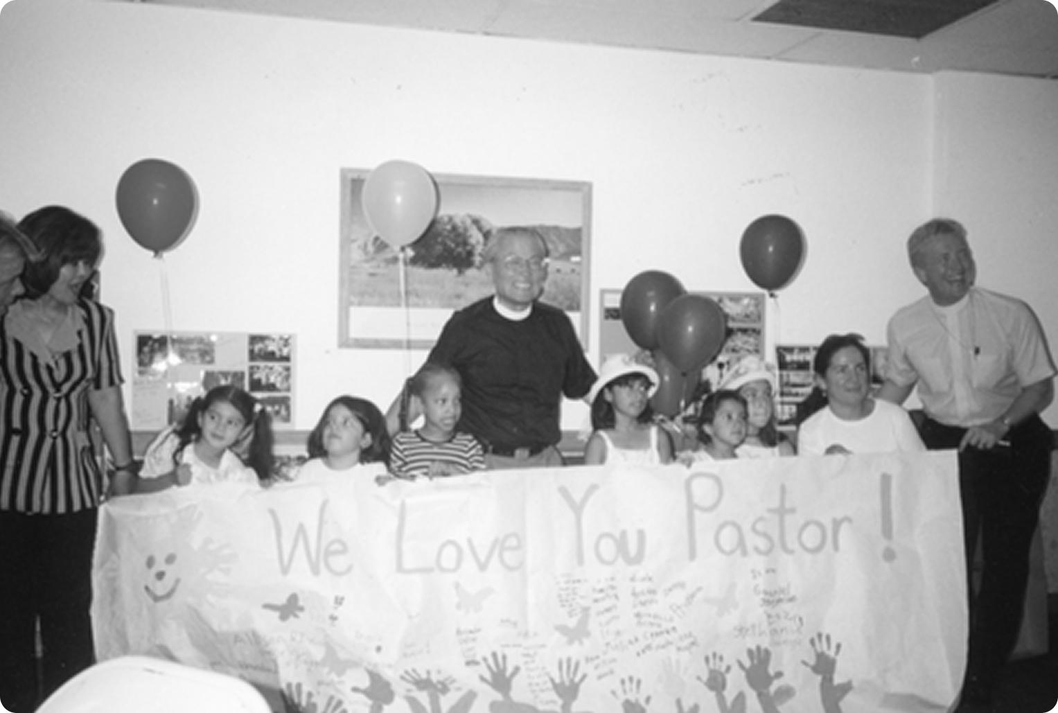 A pastor in black clerical dress smiles surrounded by young children and adults behind a table bearing a hand-painted banner reading We Love You Pastor with balloons and handprints decorating the room.