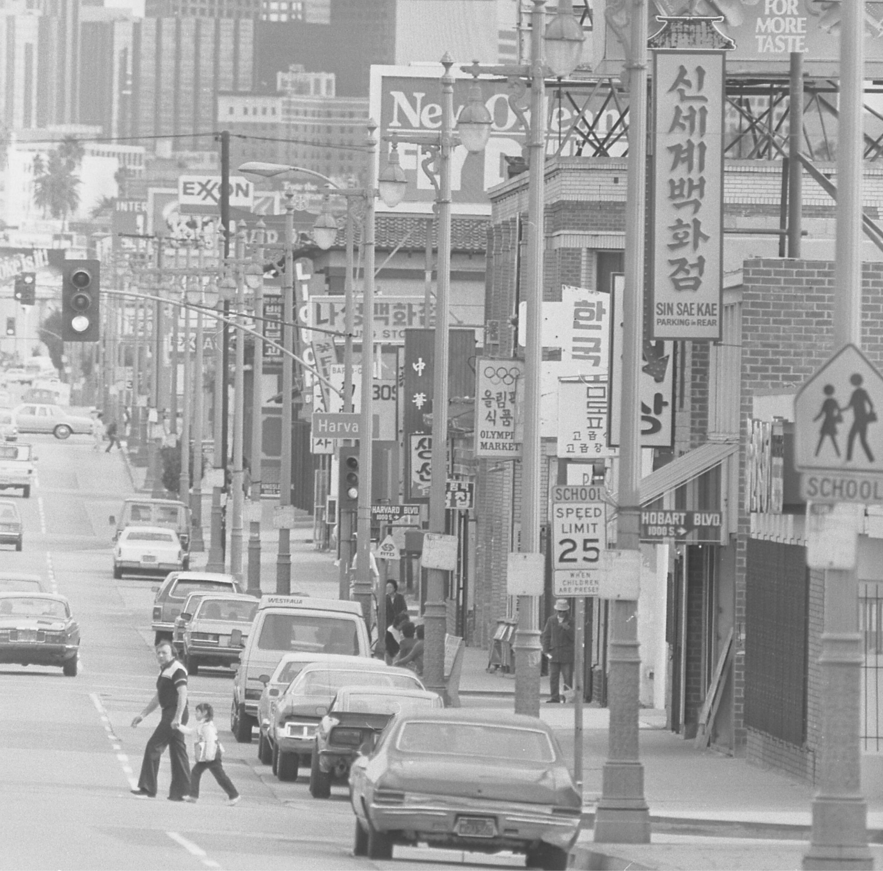 A Koreatown street in Los Angeles lined with storefronts carrying Korean-language signs, with cars parked along the curb and a pedestrian crossing the road, in a black-and-white photograph.