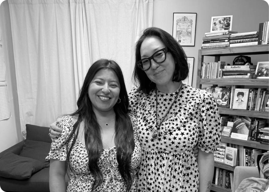 Katherine Kim and Katherine Bahena-Benitez in floral and star-print dresses smile together inside a room with a bookshelf filled with art and history books visible in the background.